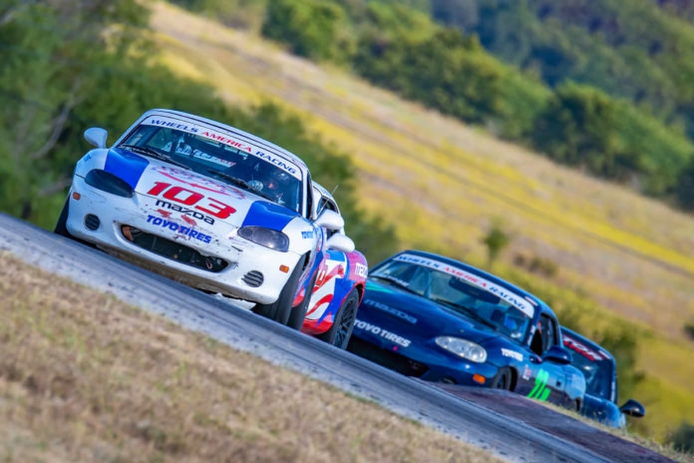 Three Spec Miatas nose to tail cresting a rise at Motorsport Ranch, Cresson
